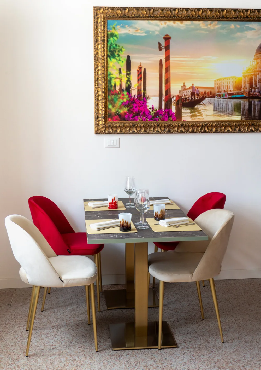 Tavolo per quattro in marmo scuro con sedie in velluto rosso e crema, sotto un quadro in cornice dorata con una veduta di Venezia.